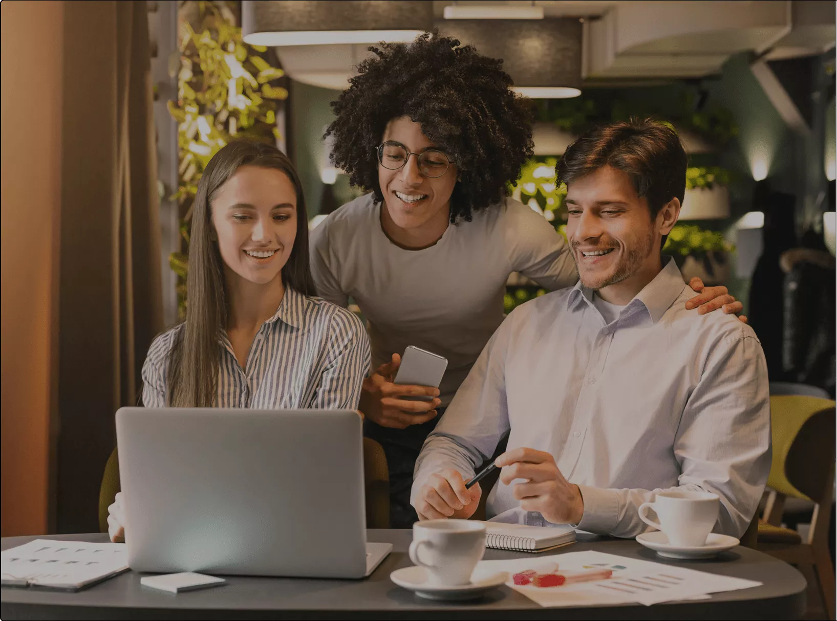 Três colegas de trabalho colaborando em ambiente moderno, com um laptop aberto e anotações na mesa. A imagem reflete um ambiente de produtividade, comunicação otimizada e trabalho em equipe.